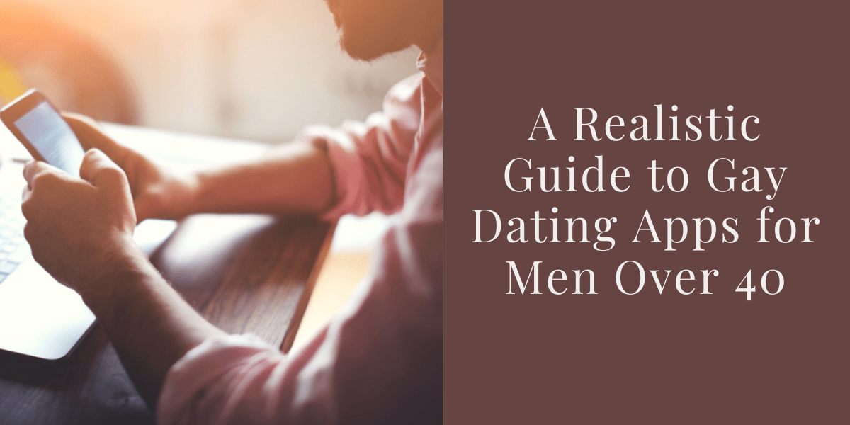 A person, presumed to be a man, has his elbows on a table that has an open laptop on it. In his hands he&#39;s holding a smartphone, that is on but the screen is difficult to see. The man is wearing a pink shirt with sleeves rolled up. We see only the child of the man, which is stubbly. The text reads &quot;A realistic guide to gay daring apps for men over 40&quot;