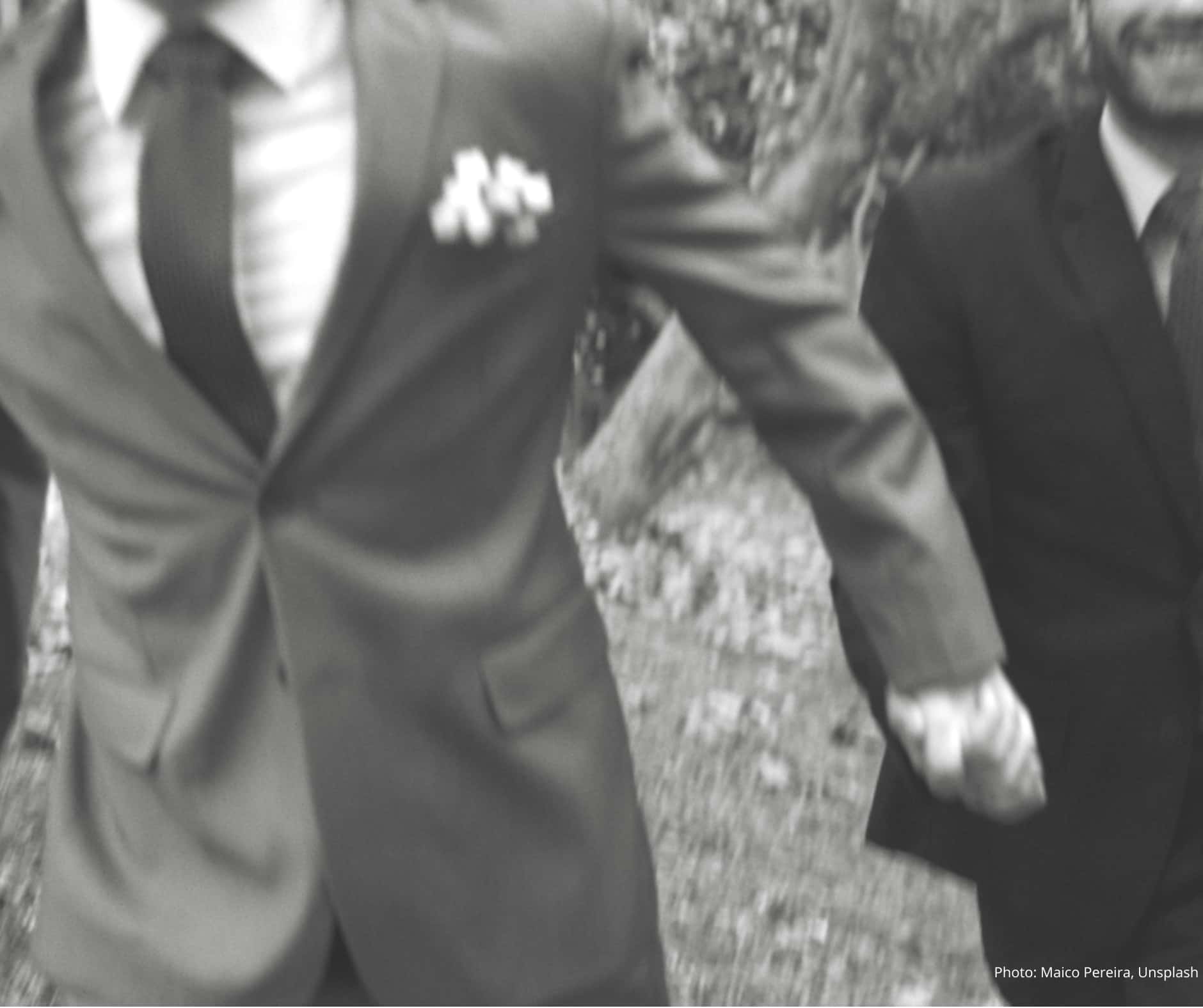 Two men, in smart wedding attire holding hands running towards the camera. Only the torso of the man on the left is visible, with a portion of the man on the right's face showing he has a beaming smile. They are outside. The photo is a black and white candid shot.