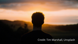 Silhouette of a person with short hair standing outdoors, facing a sunset over a landscape, with the sky glowing orange and clouds above. Credit: Tim Marshall, Unsplash.