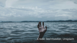 A single hand reaches out of the water in a large body of water under a cloudy sky, suggesting a person struggling or calling for help. Distant shoreline is visible in the background. Credit: Stormseeker, Unsplash.