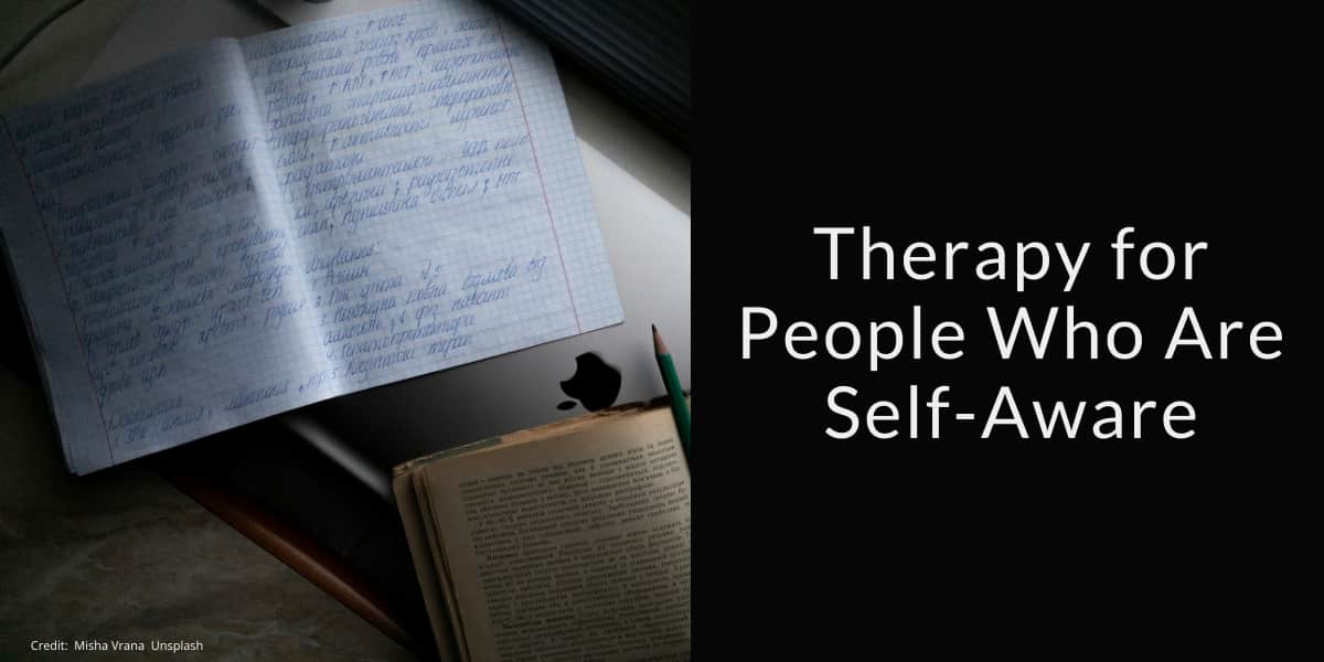 Open notebook with handwritten text in blue ink, a pencil, and an open book placed on a laptop, all on a desk. The left side of the image shows these items in soft light, while the right side features a black background with the white text: &quot;Therapy for People Who Are Self-Aware.&quot; The photo credit in small text reads: &quot;Credit: Misha Vrana, Unsplash