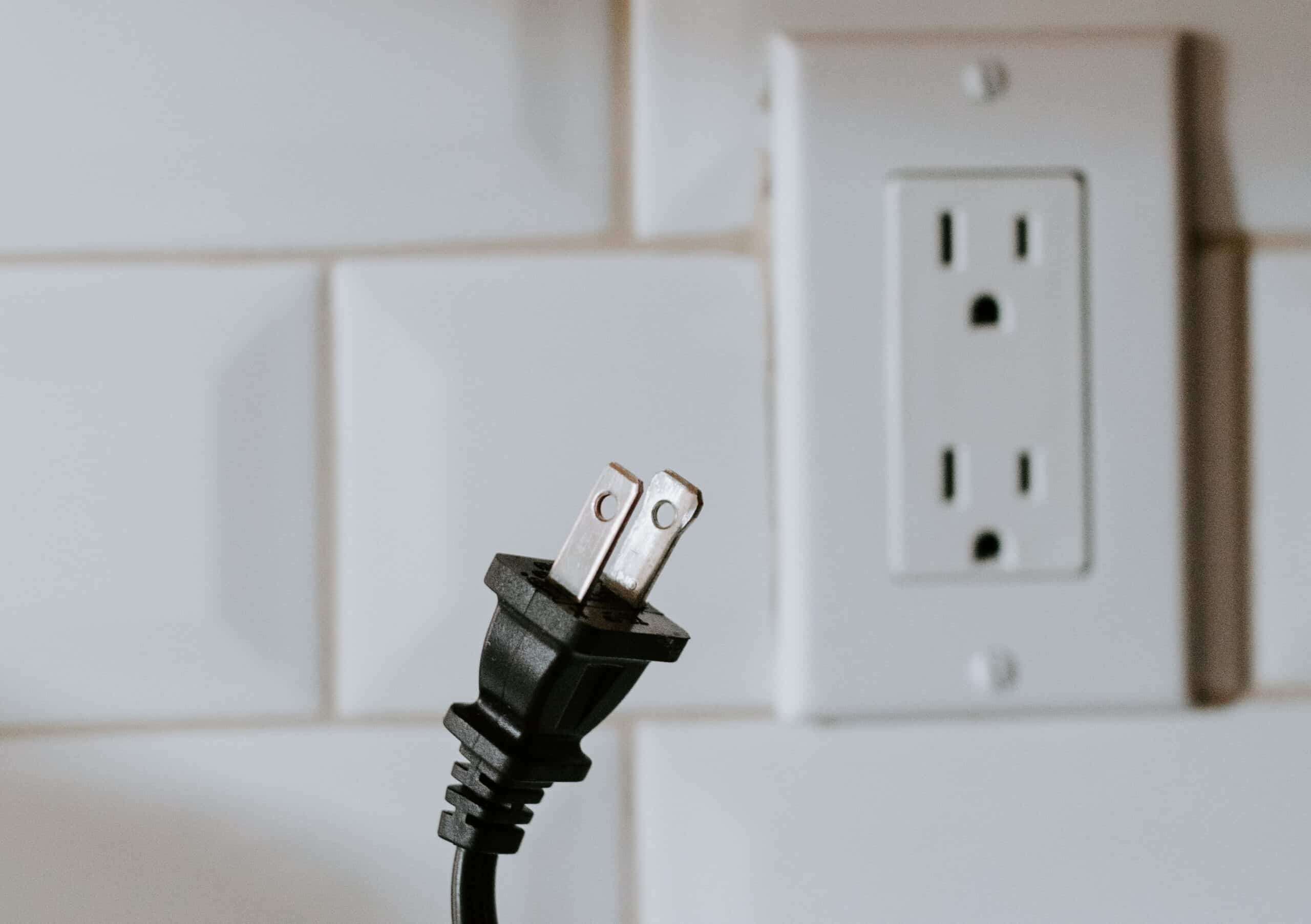 A black electrical plug, probably from an appliance in the USA, is positioned in front of a North American electrical wall plug socket.