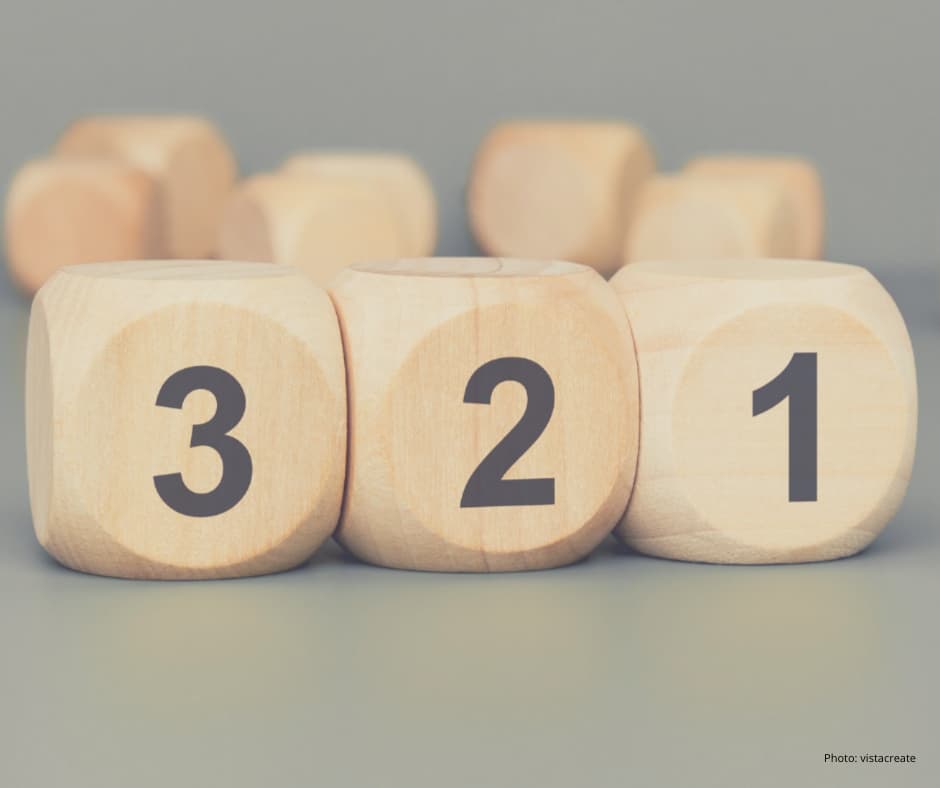 Three wooden blocks are placed side by side, each displaying a large black number: 3, 2, and 1, from left to right. The background is blurred, showing more wooden blocks out of focus. The overall tone is soft and neutral.