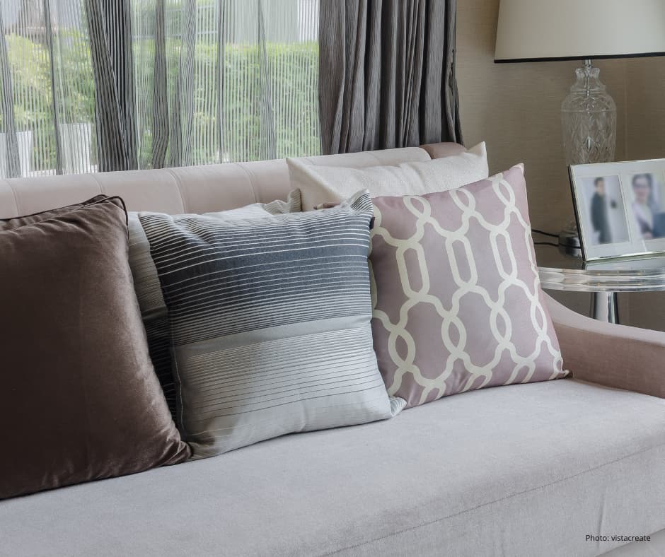 A living room with a grey sofa and a number of beige, grey and light coloured cushions arranged neatly on it. A window in the background covered by thin net curtains with a grey curtain at one side. There is a tall lamp to the right of the curtain and in front of the lamp a small table with a photo frame and two blurred photos of people in it.