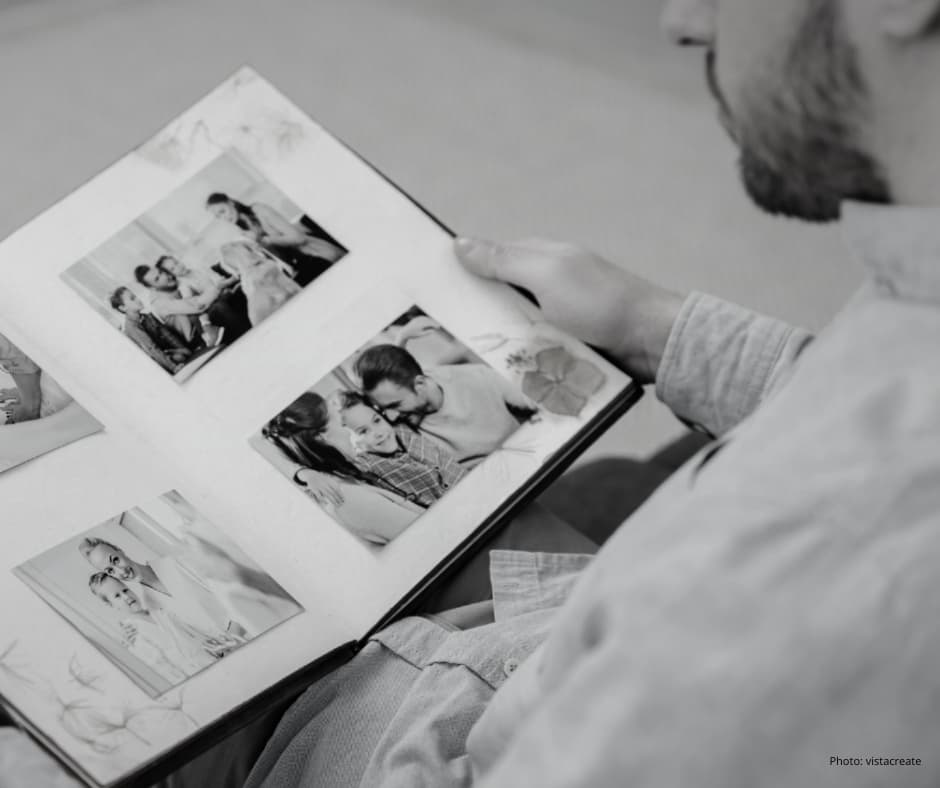 A man has a photo album on his lap. It is open on the page of four photos that depict happy family scenes of a woman man and child together.