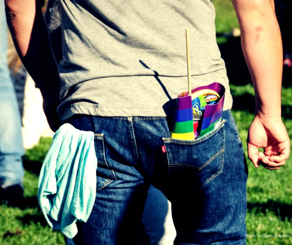 A man faces away from the camera in a field. THe frame is restricted to his lower back down to his knees. He is wearing a grey t shirt and blue Levi jeans. In the right pocked is a rainbow flag, in the left pocket is a light blue item of clothing hanging out.