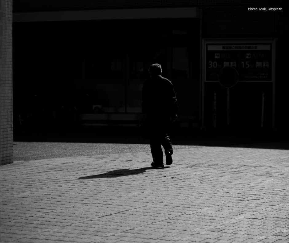 Black-and-white photograph of an older man in silhouette walking alone across a brick-paved courtyard, casting a long shadow, with a dark doorway behind him.