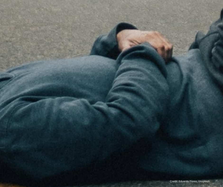 A person wearing a blue hoodie is lying on their side on a concrete surface, with their hands resting on their stomach. Only the torso and arms are visible.