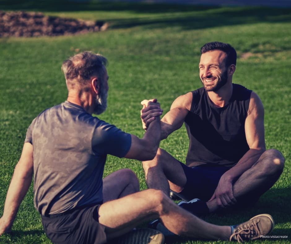 Two men sitting on grass in a park, smiling and clasping hands in a supportive gesture. One man has grey hair and a beard, the other has dark hair and a trimmed beard. They appear relaxed and friendly.