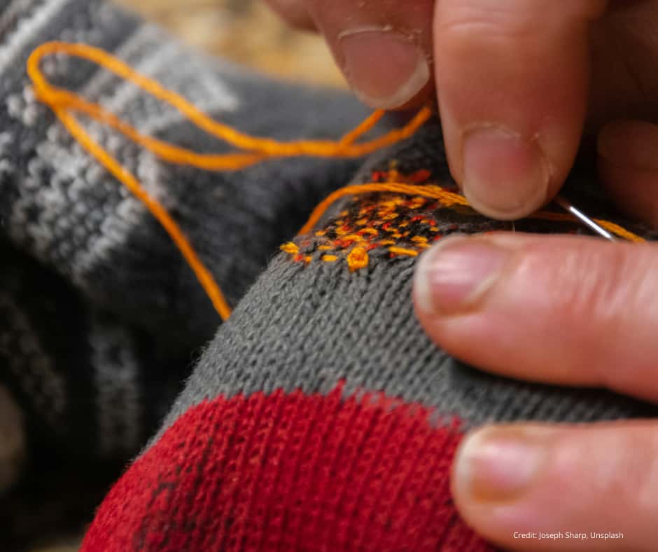 repairing hole in knitted jumper Close-up of a person’s hands mending a hole in a gray and red knitted sock using a needle and bright orange thread. The person’s fingers are holding the fabric taut while stitching, and the orange thread is visible looping through the material. The background is softly blurred, focusing attention on the repair work. Photo credit: Joseph Sharp, Unsplash.