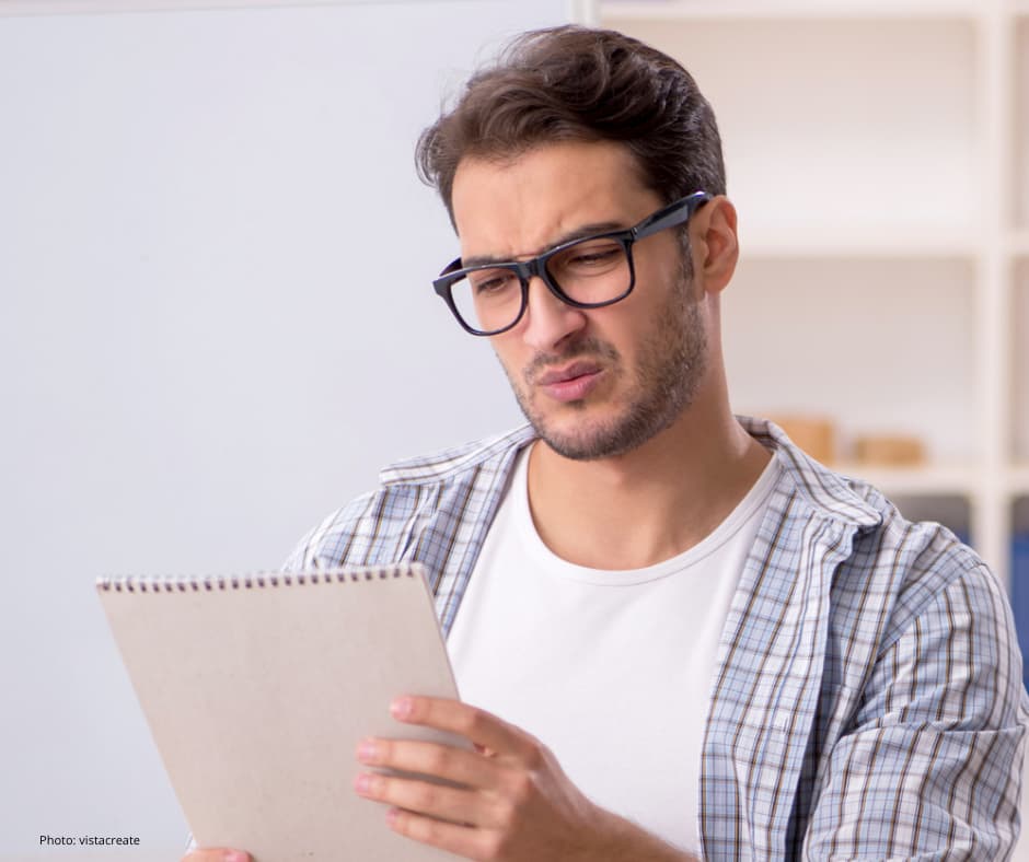 A person with short brown hair and a trimmed beard is sitting indoors, wearing a white t-shirt layered with a light blue plaid shirt. They are holding a spiral-bound notepad with both hands and looking at it. The background features shelves with books and boxes, giving the setting a casual, home or office atmosphere.