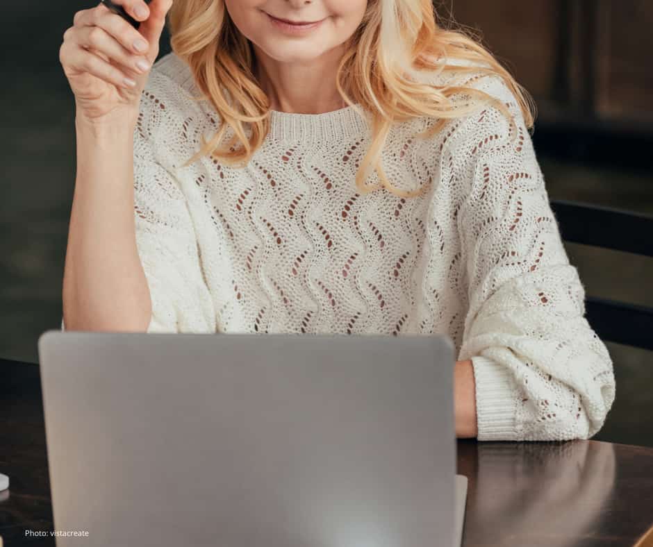 A woman with wavy blonde hair is sitting at a table, partially smiling, and looking at a laptop. She is wearing a white, textured knit sweater with a wavy pattern. Her left hand is raised, holding a pen, while her right arm rests on the table. The background is softly blurred, and the overall setting appears to be indoors.
