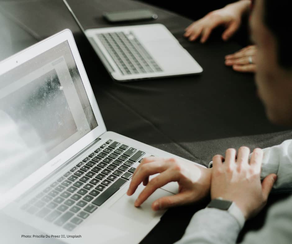 Person using a laptop computer at a table, with another laptop and hands visible in the background, suggesting a collaborative or work setting.
