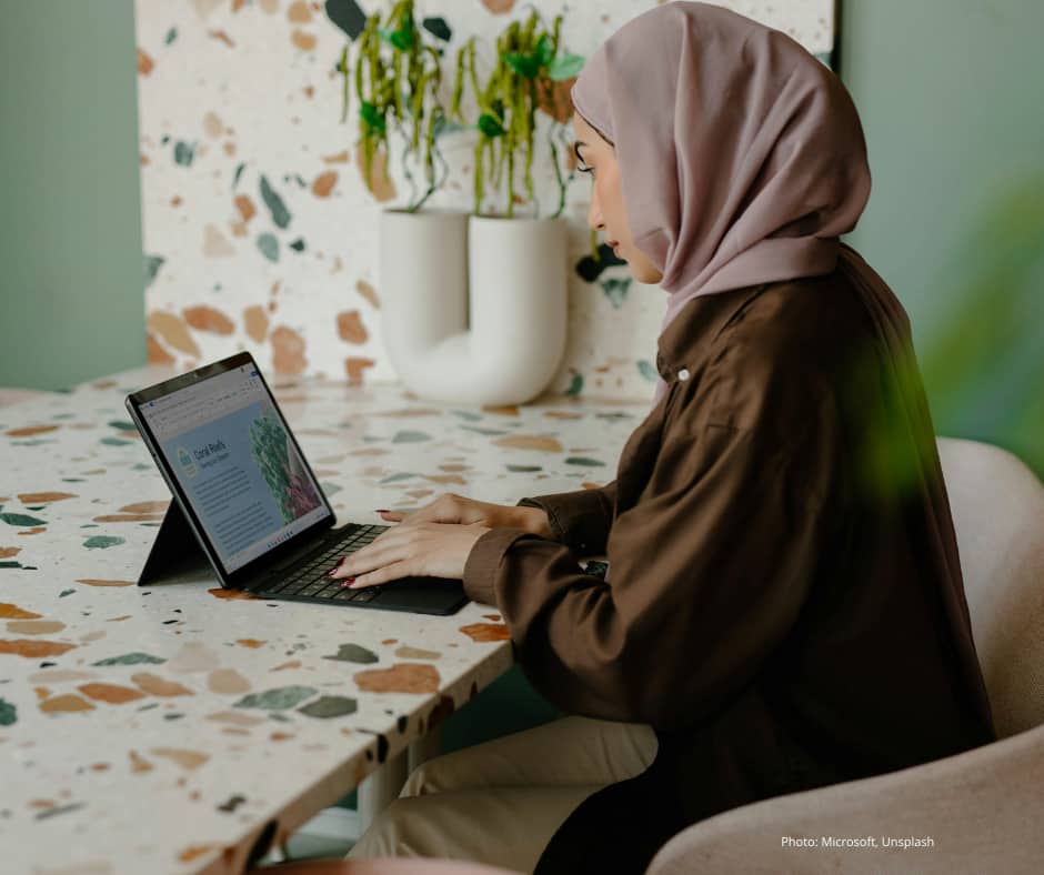 A woman wearing a light pink hijab and brown shirt is sitting at a terrazzo-patterned table, working on a tablet with a keyboard. The background features a decorative plant and a light green wall. The setting appears modern and calm, suggesting a comfortable workspace.