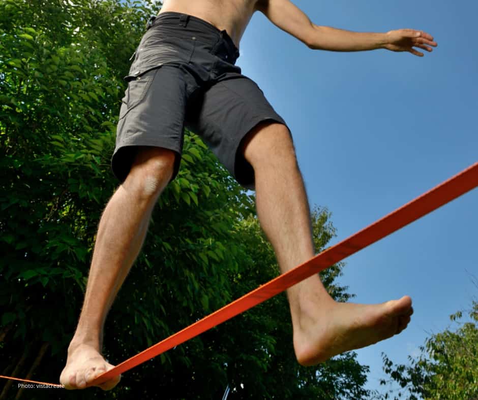 A person wearing black shorts is balancing barefoot on a slackline outdoors, with trees in the background and a clear blue sky above. The image is taken from a low angle, focusing on the legs and feet.